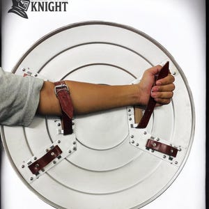 May include: A silver-colored, round shield with a handle and arm straps. The shield has a riveted design and a brown leather handle and straps. The text "MEDIVAL KNIGHT" is in the upper left corner.