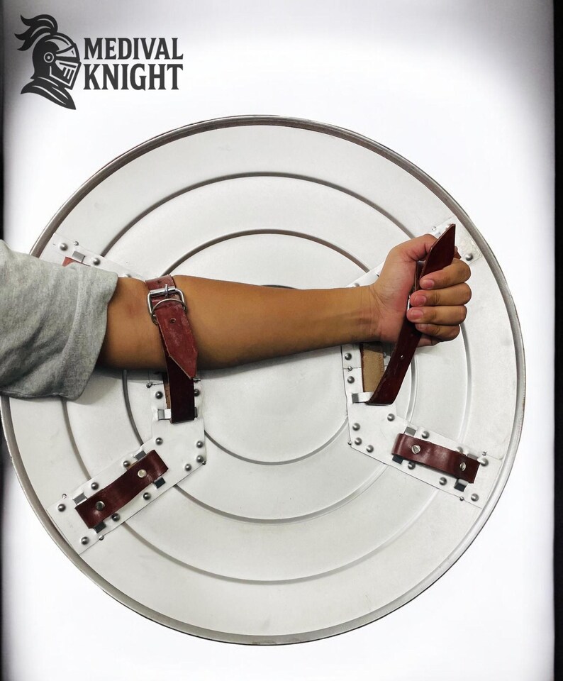 May include: A silver-colored, round shield with a handle and arm straps. The shield has a riveted design and a brown leather handle and straps. The text "MEDIVAL KNIGHT" is in the upper left corner.