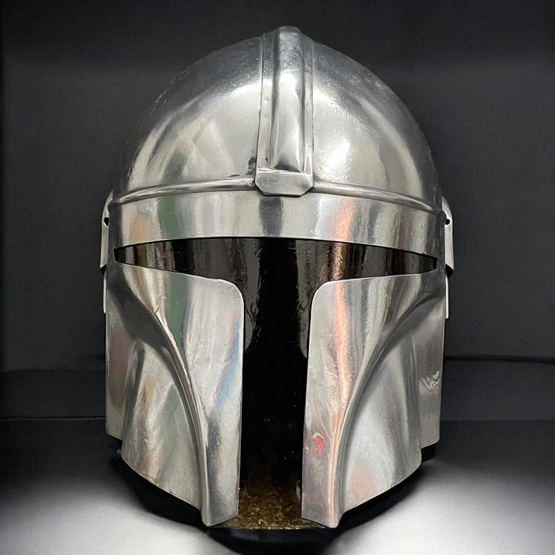 May include: A metallic silver Mandalorian helmet with a distinctive T-shaped visor. The helmet has a smooth, reflective surface and a prominent central ridge. The design is reminiscent of science fiction and space opera themes.