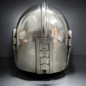 May include: A silver-colored helmet with a rounded top and a series of rectangular and vertical design elements. The helmet has a smooth, reflective surface and appears to be made of metal. The back of the helmet features a series of rectangular panels.