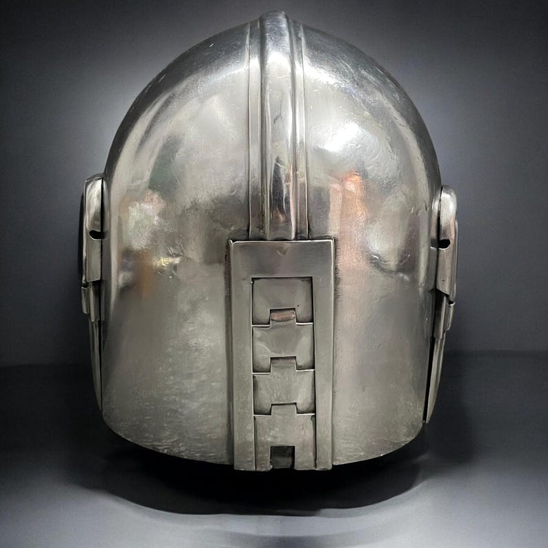 May include: A silver-colored helmet with a rounded top and a series of rectangular and vertical design elements. The helmet has a smooth, reflective surface and appears to be made of metal. The back of the helmet features a series of rectangular panels.