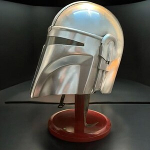 May include: A silver-colored Mandalorian helmet, displayed on a red stand. The helmet has a visor and detailed side features. The stand is round with a curved base, set against a dark background.