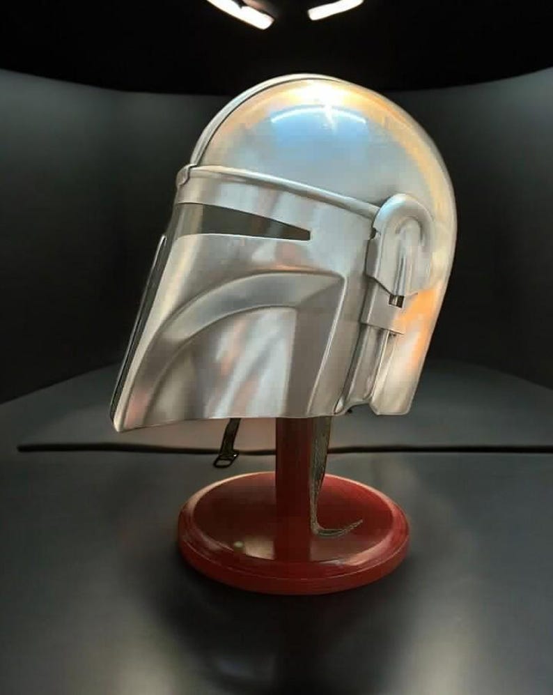 May include: A silver-colored Mandalorian helmet, displayed on a red stand. The helmet has a visor and detailed side features. The stand is round with a curved base, set against a dark background.