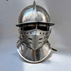 May include: A polished, silver-colored medieval knight's helmet. The helmet features a visor with horizontal slits for vision and ventilation, a pointed top, and a neck guard. The logo "MEDIVAL KNIGHT" is visible in the upper left corner.