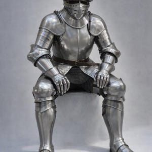 May include: Full suit of polished steel armor, including a helmet with a visor, shoulder and arm protection, and leg and foot coverings. The armor is complete with a chainmail shirt and a brown belt. The knight is seated.