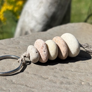 May include: A keychain featuring five smooth, rounded stones in shades of cream, beige, and light pink, strung together on a piece of natural twine and attached to a circular silver-toned metal ring, with frayed twine ends extending from the last stone.