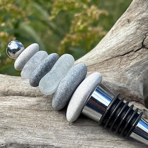 May include: A bottle stopper featuring a stack of smooth, rounded gray and white stones topped with a polished silver-tone metal sphere. The base of the stopper is made of shiny chrome-finished metal with black rubber rings for a secure fit.