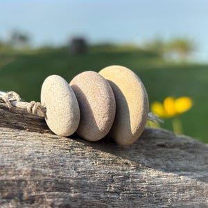 May include: A keychain featuring three smooth, rounded, light beige stones of varying sizes strung together on a piece of natural-colored twine. The stones are arranged in a row, attached to a circular silver-toned metal ring at one end.