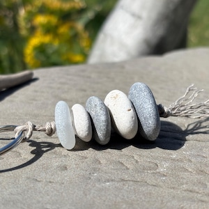 May include: A keychain featuring a silver metal ring attached to a piece of light brown string. The string holds five smooth, rounded stones of varying sizes and colors, including a translucent white piece, a small white stone, and three grey stones.