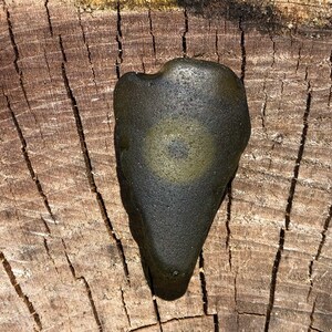 May include: A dark green, heart-shaped stone with a light green circle in the center. The stone is sitting on a wooden surface.