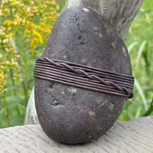 May include: A smooth, dark brown stone with subtle green speckles, wrapped horizontally with multiple strands of thin, dark brown leather cord that are twisted together at the center to create a decorative band around the center of the stone.