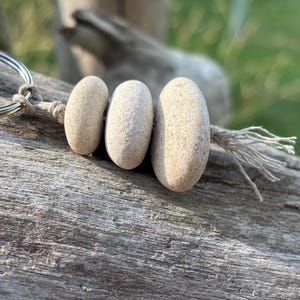 May include: A keychain featuring three smooth, light beige oval stones strung together on a piece of natural-colored twine. The stones are attached to a circular silver-toned metal split ring, and the twine ends in a small, frayed tassel.