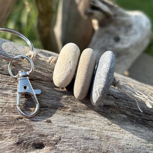 May include: A keychain featuring a silver-toned metal split ring and a lobster clasp attached to a piece of twine. Three smooth, oval-shaped stones in shades of beige, tan, and grey are threaded onto the twine, ending in a small knotted tassel.