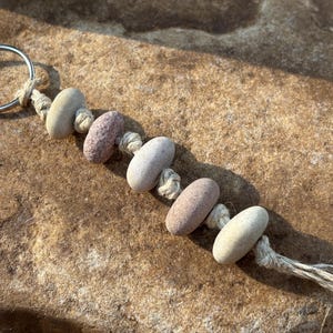 May include: A keychain featuring five smooth, oval-shaped stones in shades of beige, tan, and dusty rose, strung together with natural-colored twine. The stones are separated by knots in the twine, and a silver-toned metal ring is attached at the top.