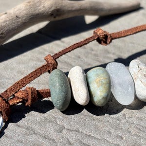 May include: A keychain featuring five smooth, rounded stones in shades of sage green, off-white, mottled green, translucent white, and speckled gray. The stones are strung onto a brown leather cord with knots, attached to a silver metal ring.
