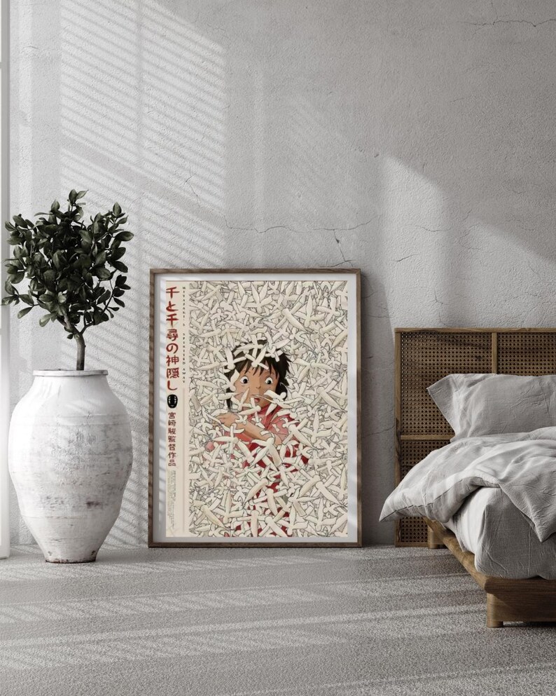 May include: A framed poster featuring a character from the animated film "Spirited Away." The artwork depicts a young girl surrounded by white objects, with Japanese text in the upper left corner. The poster is displayed in a room with a plant and a bed.