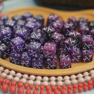 May include: A collection of purple and black dice with white numbering, arranged on a wooden tray. The dice are surrounded by decorative beads in shades of pink and red, creating a visually appealing arrangement. The dice are for tabletop games.