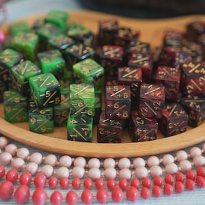 May include: A wooden tray holds a collection of dice in shades of green and burgundy, with gold markings. The dice are arranged in a pile, suggesting a game or hobby. Pink and red beads are in the foreground.