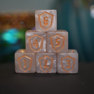 May include: Six pearlescent dice with gold-colored shield designs and numbers 1 through 6 stacked in a pyramid. The dice are on a dark surface with a blurred background of gold and teal.