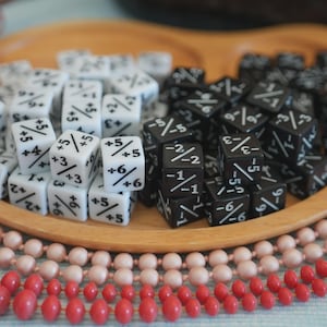 Puede incluir: Bandeja de madera con dos secciones llenas de dados. Una sección tiene dados blancos con marcas negras, la otra dados negros con marcas blancas. Collares de cuentas rosas y rojas y bayas decorativas rodean la bandeja.