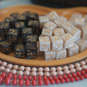 May include: A wooden tray holds two sets of dice. One set is black with gold markings, the other is white with gold markings. The dice are arranged in neat piles. Pink and red beads surround the tray.