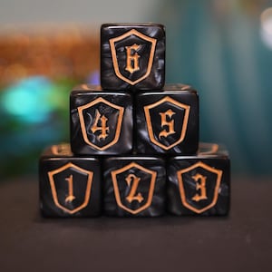 May include: A stack of six black dice with gold-colored numbers and shield designs. The dice are arranged in a pyramid shape, with numbers 1 through 6 visible. The dice are made of a marbled material.