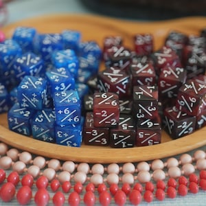 Op de afbeelding: Een houten dienblad bevat twee sets dobbelstenen, één blauw en één bordeaux, met witte cijfers. De dobbelstenen zijn omgeven door roze en rode kralenkettingen en roze bessen. De dobbelstenen zijn voor tabletopspellen.