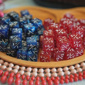 May include: Wooden tray holding two piles of dice. One pile is blue with white markings, the other is red with white markings. Decorative beads in pink and red surround the tray. Dice are used for tabletop games.