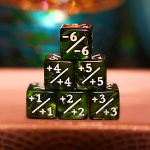 May include: A stack of six green dice with white markings. Each die has a different combination of numbers and symbols, including +1, +2, +3, +4, +5, and -6. The dice are arranged in a pyramid shape, with the top die showing -6.
