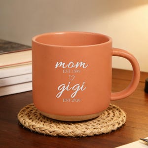 May include: A peach-coloured ceramic mug with the words "mom" and "gigi" in white script. The mug sits on a round, woven coaster. The mug has a large handle and the text "EST. 1989" and "EST. 2026".