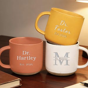 Puede incluir: Una taza de cerámica blanca con un asa negra y el texto "But First, Coffee" en negrita negra. La taza se encuentra sobre un fondo borroso, sugiriendo un ambiente acogedor.