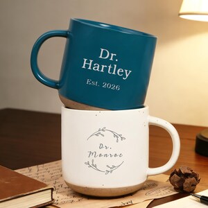 Puede incluir: Dos tazas de cerámica apiladas. La superior es de color turquesa con el texto blanco "Dr. Hartley Est. 2026". La inferior es blanca con "Dr. Monroe" dentro de una corona floral. Bases naturales.
