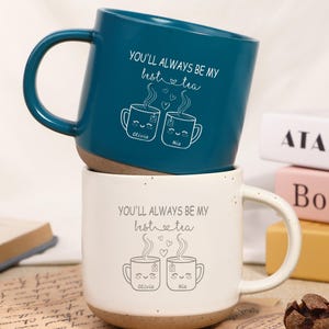 May include: Two ceramic mugs, one teal and one white, stacked. The teal mug has a black handle and the text "YOU'LL ALWAYS BE MY hot tea" with two cartoon tea cups. The white mug has the same text and design.