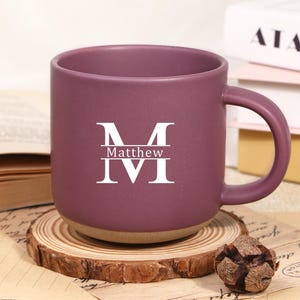 May include: A plum-coloured ceramic mug with a white monogram and the name "Matthew" printed on the front. The mug has a rounded handle and a light brown base. It sits on a wooden coaster.