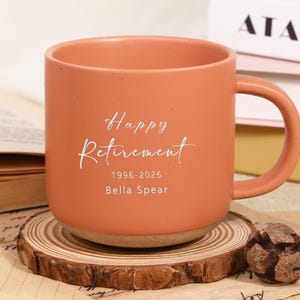 Puede incluir: Una taza de cerámica color melocotón con asa, con las palabras "Happy Retirement" y los años 1996-2026, junto con el nombre "Bella Spear" en escritura blanca. La taza está sobre un posavasos de madera.