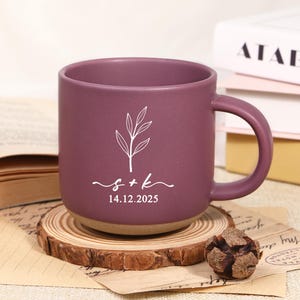 Peut inclure: Une tasse en céramique de couleur prune avec un motif floral blanc et les initiales "s + k" avec la date "14.12.2025" imprimée en dessous. La tasse a une forme arrondie et une base beige. Elle repose sur un dessous de verre en bois.