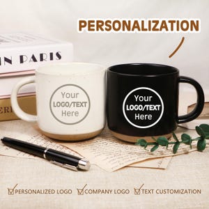 May include: Two ceramic mugs, one white and one black, with a customizable logo area. The mugs have a speckled finish and a natural-coloured base. A black pen rests in front of the mugs. Text on the image reads "PERSONALIZATION".