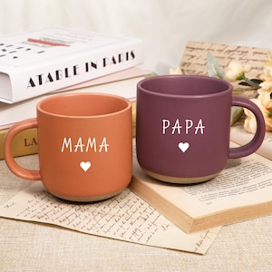 Puede incluir: Dos tazas de cerámica, una naranja y otra morada, cada una con una palabra blanca y un corazón. La taza naranja dice "MAMA", y la morada dice "PAPA". Ambas tazas tienen una base de color neutro y un asa.