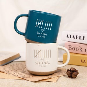 May include: Two ceramic mugs, one teal and one white, stacked. The teal mug has the text "Leo & Mia 12.12.2016". The white mug has the text "Jack & Chloe 12.11.2016". Both mugs have a tally mark design and a heart.