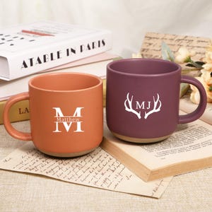 Könnte beinhalten: Zwei Keramikbecher, einer orangefarben und einer pflaumenfarben, jeweils mit einem weißen Monogramm. Der orangefarbene Becher hat den Namen "Matthew" und die Initialen "M IVI". Der pflaumenfarbene Becher zeigt die Initialen "MJ" mit einem Hirschgeweih-Design.
