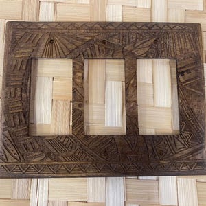 May include: A rectangular, dark brown wooden light switch cover with three openings. The cover is intricately carved with geometric and stylized leaf patterns. The background is a woven, light-colored material.