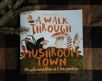 May include: A children's book titled "A Walk Through Mushroom Town" by Nicole and David Charpentier. The cover features a white background with colorful illustrations of mushrooms and a forest scene.