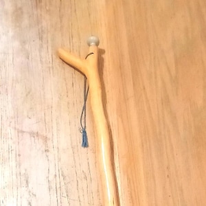 May include: A light-colored wooden walking stick with a rounded top and a small blue tassel. The stick has a natural, organic shape and a white tip. The walking stick is made of wood.