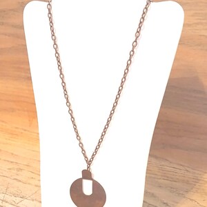 May include: A copper-coloured necklace with a long chain and a pendant. The pendant is a circular shape with a rectangular top and a U-shaped cutout in the centre. The necklace is displayed on a white mannequin.