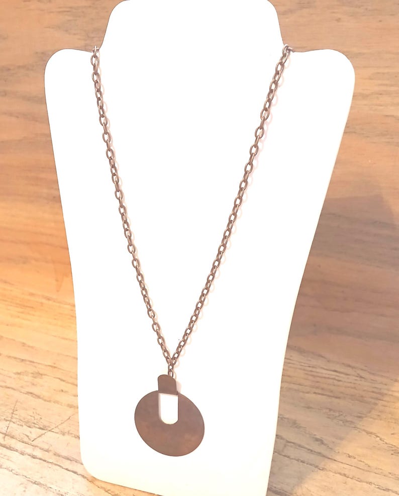 May include: A copper-coloured necklace with a long chain and a pendant. The pendant is a circular shape with a rectangular top and a U-shaped cutout in the centre. The necklace is displayed on a white mannequin.
