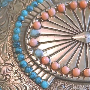May include: Close-up of a silver-toned belt buckle with intricate floral scrollwork. The buckle is adorned with turquoise and coral-colored beads, arranged in a decorative pattern. The design has a vintage, Western aesthetic.