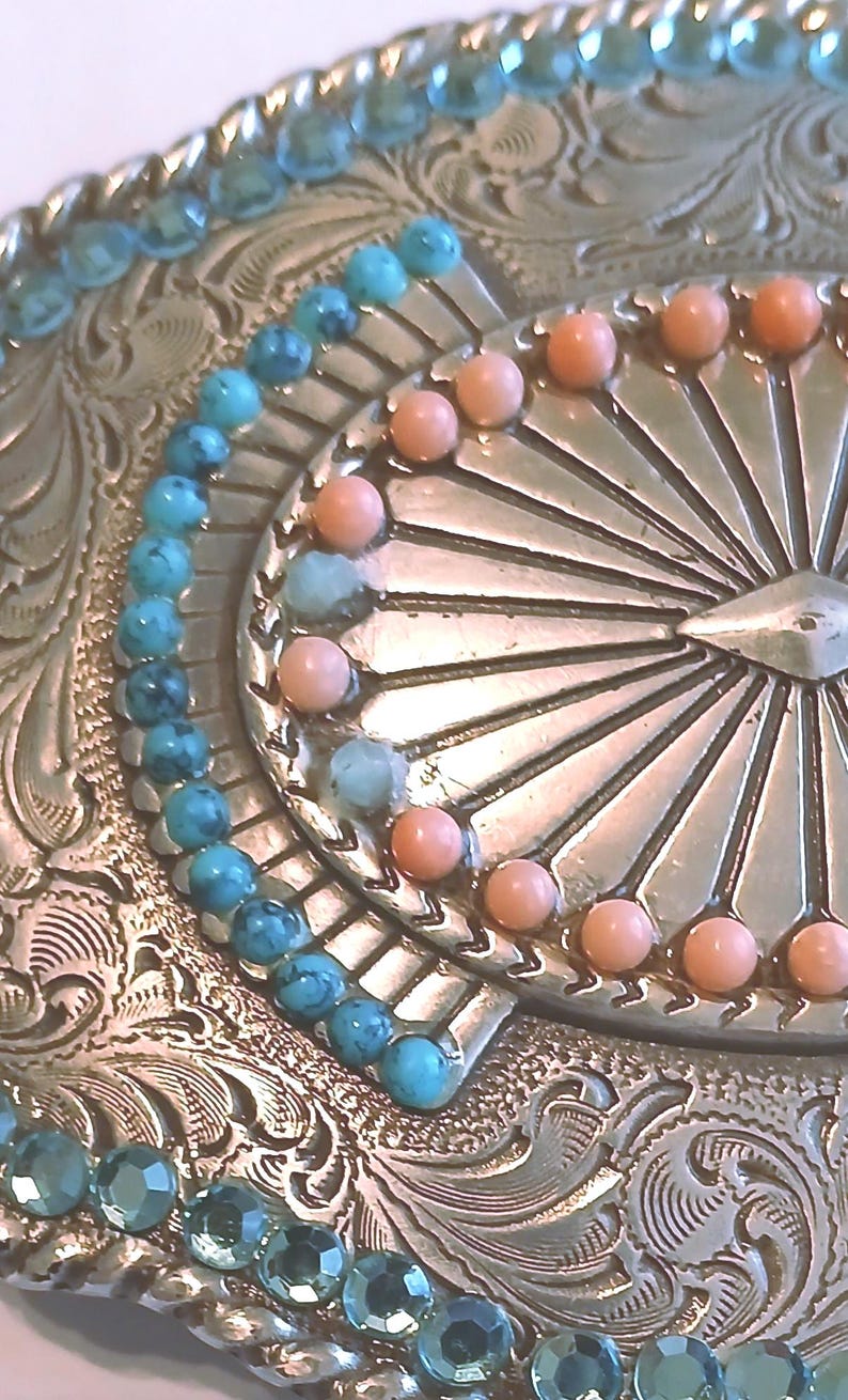 May include: Close-up of a silver-toned belt buckle with intricate floral scrollwork. The buckle is adorned with turquoise and coral-colored beads, arranged in a decorative pattern. The design has a vintage, Western aesthetic.