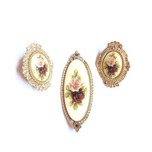 May include: Three vintage brooches with floral designs. The brooches feature oval and oblong shapes with cream-colored backgrounds and painted roses in shades of pink and burgundy. The brooches are framed in gold-toned metal with decorative detailing and rhinestones.