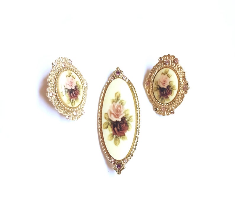 May include: Three vintage brooches with floral designs. The brooches feature oval and oblong shapes with cream-colored backgrounds and painted roses in shades of pink and burgundy. The brooches are framed in gold-toned metal with decorative detailing and rhinestones.