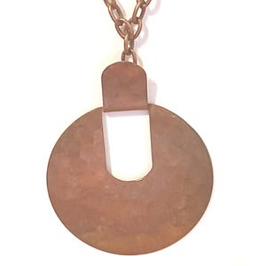 May include: A copper-coloured pendant necklace. The pendant features a circular design with a rectangular top and a U-shaped cutout. The chain is made of matching copper-coloured links. The necklace is set against a white background.